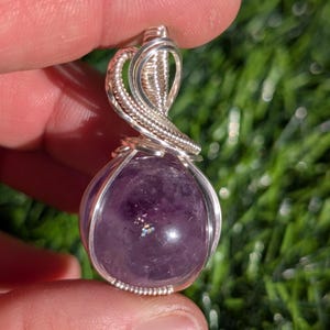 Può includere: Un ciondolo in ametista rotonda di colore viola intenso, racchiuso in una montatura decorativa in filo d'argento. La lavorazione del filo presenta bobine e anelli complessi che formano una gabbia sicura attorno alla pietra, estendendosi verso l'alto.