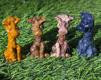 May include: Four carved giraffe figurines in shades of orange, brown, and blue sit on green grass. Each giraffe is in a seated position with detailed features. The figurines vary in colour and pattern, showcasing the natural stone.