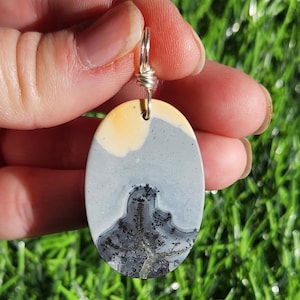 May include: Oval pendant with a landscape design. The pendant features a silver wire bail and a natural stone with a yellow, gray, and black landscape pattern. The stone is held by a silver wire. The background is green grass.