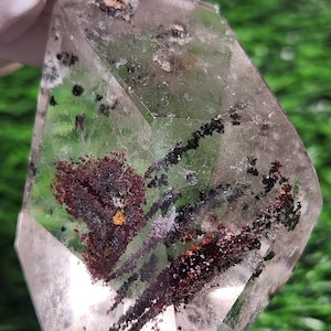 Puede incluir: Un cristal de cuarzo transparente con inclusiones internas de minerales rojo oscuro, negro y amarillo. El cristal se sostiene en una mano enguantada, con un fondo verde visible.