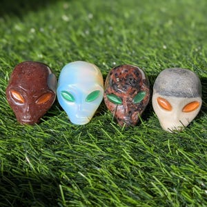 May include: Four alien head sculptures in various colors and textures. One is reddish-brown with orange eyes, another is light blue with green eyes, a third is speckled red and black with green eyes, and the last is gray with orange eyes.