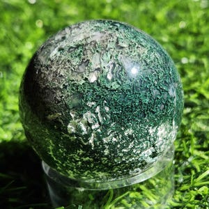 May include: A polished moss agate sphere featuring intricate, deep green dendritic inclusions that resemble organic foliage patterns against a translucent, milky white and grey crystalline background, resting on a clear circular acrylic stand.