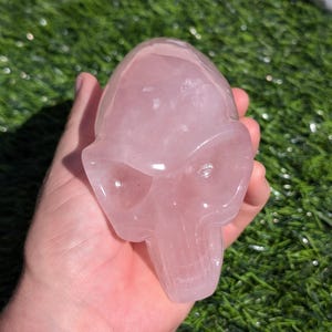 Pode incluir: Uma escultura de crânio de quartzo rosa. O crânio é esculpido com detalhes precisos, incluindo órbitas oculares, um nariz e uma boca.