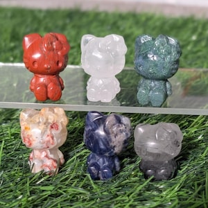 Puede incluir: Seis figuras de gato de piedras preciosas en varios colores: rojo, transparente, verde, beige, azul y gris. Las figuras están expuestas sobre una estantería transparente sobre un fondo de hierba verde.
