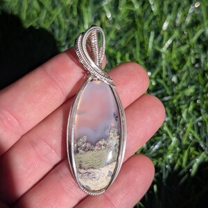 May include: An oval-shaped pendant featuring a translucent, milky stone with mossy green and earthy brown inclusions, encased in a polished silver wire-wrapped setting with a decorative coiled bail at the top, showcasing intricate metalwork.