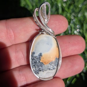 May include: An oval-shaped pendant featuring a smooth, multi-colored stone with sections of white, soft orange, and dark gray speckled patterns. The stone is encased in a decorative silver wire-wrapped setting with a coiled loop at the top.