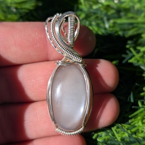 May include: A wire-wrapped pendant featuring a smooth, oval-shaped, translucent gray stone encased in polished silver-colored metal wire. The wire forms an intricate, coiled bail at the top, securely holding the stone in a vertical orientation.