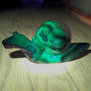 Puede incluir: Figura de caracol de malaquita verde y negra. La concha del caracol tiene un patrón arremolinado de verde oscuro y negro, mientras que el cuerpo es de un verde brillante. La figura está sobre una superficie de madera.