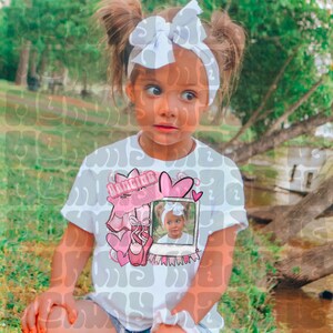 May include: A young girl wearing a white t-shirt with a pink and white graphic design featuring the word "Dancing", a ballet shoe, and a photo. She has a white bow headband and her hair is in pigtails.