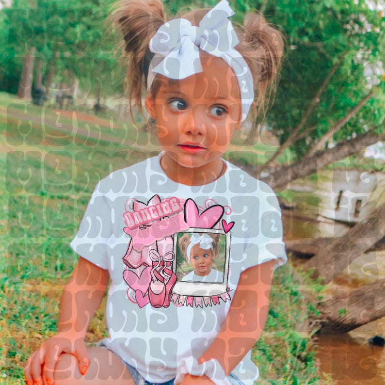 May include: A young girl wearing a white t-shirt with a pink and white graphic design featuring the word "Dancing", a ballet shoe, and a photo. She has a white bow headband and her hair is in pigtails.