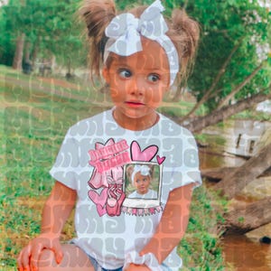 May include: A young girl wearing a white t-shirt with a pink graphic that says "Dancing Queen". The graphic includes ballet shoes, hearts, and a photo. She has a white bow headband and her hair is in pigtails.
