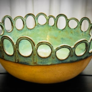 Może przedstawiać: Ręcznie wykonana ceramiczna miska o unikalnym wzorze. Miska ma jasnozielone wnętrze i kontrastującą żółto-pomarańczową podstawę. Obręcz jest ozdobiona serią owalnych wycięć, dodających dekoracyjnego akcentu.