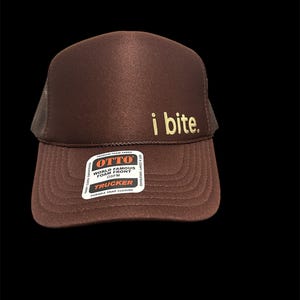 May include: A brown trucker hat with the text "i bite" in gold lettering. The hat features a foam front and a mesh back. The hat has a snap closure and a tag that reads "World Famous Foam Front".