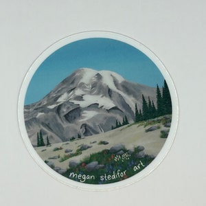 May include: A circular sticker with a watercolor painting of a mountain range with snow-capped peaks. The mountain range is set against a blue sky. The sticker has the text "megan stedifor art" written at the bottom.