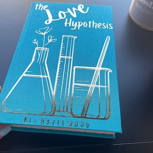 Puede incluir: Un libro de tapa dura azul turquesa con el título "The Love Hypothesis" en letras blancas. La portada presenta una ilustración plateada de un vaso de precipitados, una probeta graduada y una varilla de agitación.