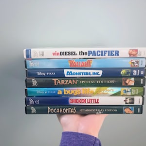 May include: A stack of eight DVDs featuring animated films, including "The Pacifier", "Valiant", "Monsters, Inc.", "Tarzan", "A Bug's Life", "Chicken Little", and "Pocahontas".