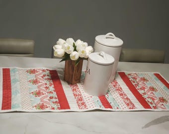 May include: A white quilted table runner with red and blue floral patterns. Two white ceramic canisters with lids are on the runner, one with the word "TEA" on it. A bouquet of white tulips in a wooden vase is on the runner.