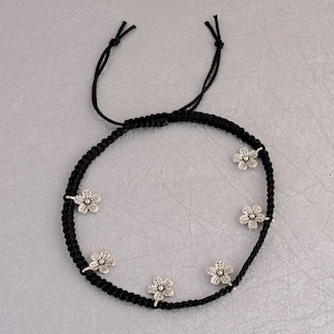May include: Black ankle bracelet with silver flower charms. The bracelet is made of a braided cord and has six flower charms.