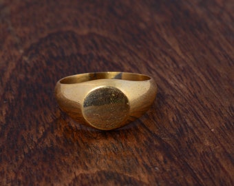 Puede incluir: Un anillo de sello de oro con una parte superior redonda y lisa. El anillo está sobre una superficie de madera marrón oscura.