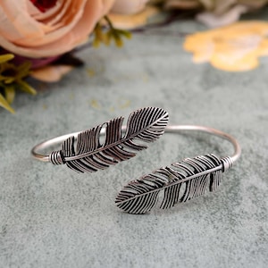 May include: Silver tone open cuff bracelet with two feather designs. The bracelet is open on one side and has a simple, minimalist design.