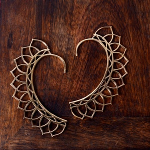 Puede incluir: Dos puños de oreja dorados con un diseño floral, sobre una superficie de madera oscura. Los puños de oreja se curvan alrededor de la oreja, con una serie de formas de pétalos a lo largo del borde exterior. El diseño es intrincado y delicado.