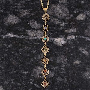 May include: A gold chain necklace featuring seven chakra symbols and gemstones. Each symbol is a mandala with a gemstone at its centre. The gemstones are purple, indigo, blue, green, yellow, orange and red.