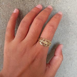 May include: A gold ring with a delicate floral design. The ring is worn on the middle finger of a hand.