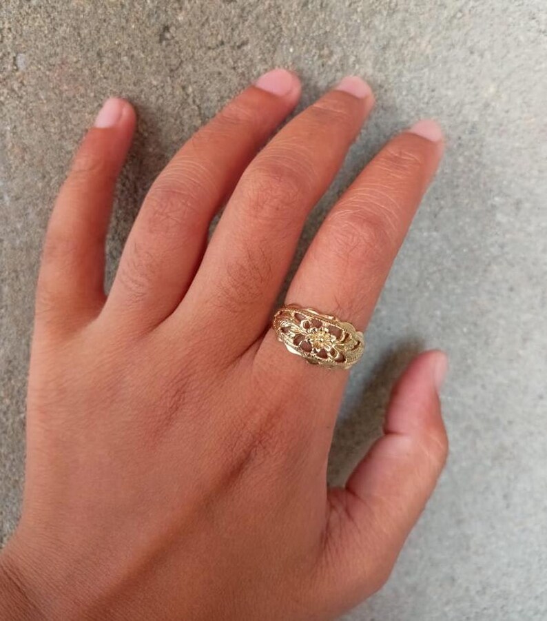 May include: A gold ring with a delicate floral design. The ring is worn on the middle finger of a hand.