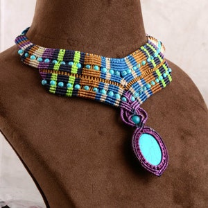May include: A handmade macrame necklace with a turquoise pendant. The necklace features a woven design in blue, purple, gold, and green, accented with turquoise beads. The pendant is an oval turquoise stone set in a purple macrame frame.