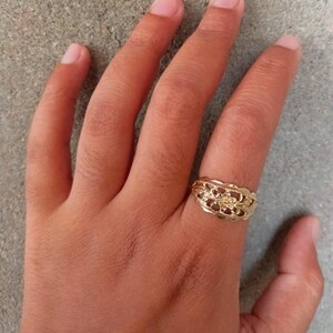 May include: A gold ring with a delicate floral design. The ring is worn on the middle finger of a hand.