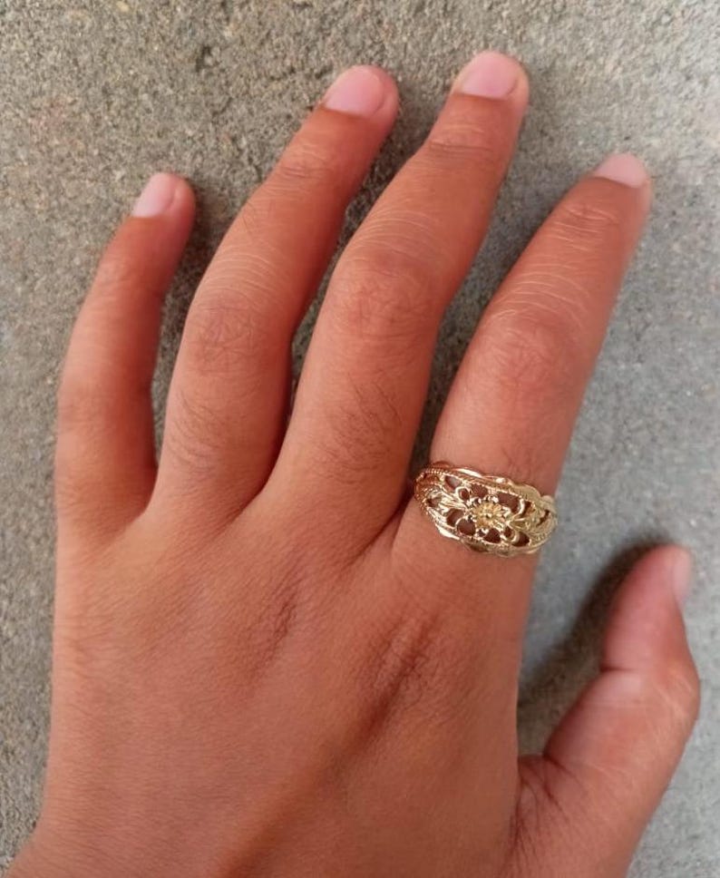 May include: A gold ring with a delicate floral design. The ring is worn on the middle finger of a hand.