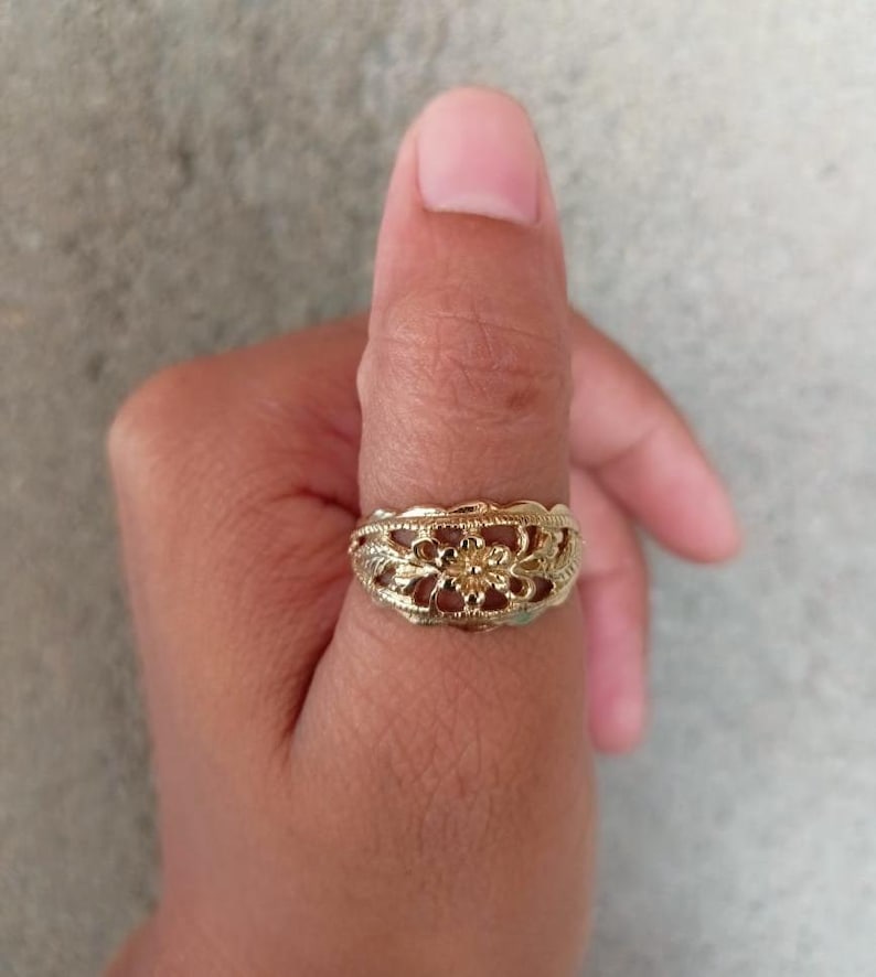 May include: A gold filigree ring with a floral design. The ring is worn on a person's thumb.