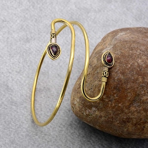 May include: A gold-colored arm cuff with a teardrop-shaped, dark red gemstone at each end. The cuff has an open design and is resting on a stone surface. The gemstones are set in a decorative, textured metal setting.