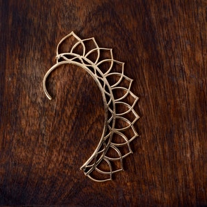 May include: A gold-colored ear cuff with a floral design. The cuff curves around the ear and features a series of petal-shaped cutouts. The jewelry is made of a thin metal and rests on a dark wood surface.