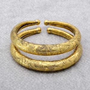 Könnte beinhalten: Zwei goldfarbene Metallarmbänder mit aufwendigen Blumenmustern. Die Armbänder sind übereinander gestapelt.