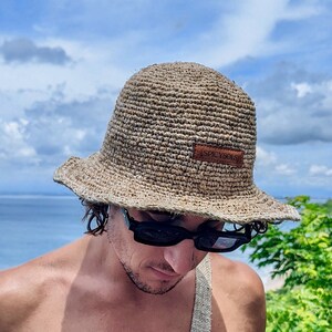 May include: A person wearing a straw hat with the text "SIMPLY" on it, sunglasses, and a beige fanny pack. The person is wearing blue swim trunks and is standing in front of a body of water.