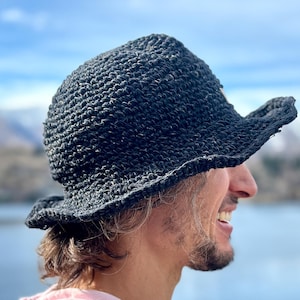May include: A person wearing a black crocheted hat with a wide brim, smiling and looking to the right. They are wearing a pink hoodie and are standing in front of a body of water.