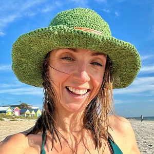 May include: A woman wearing a green crochet bikini top and a green crochet sun hat. She is smiling and has a silver necklace with a rectangular pendant.