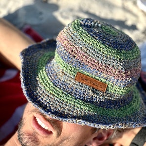 May include: A multi-coloured, crocheted bucket hat with a brown leather patch that reads "SPICYSOLS". The hat features shades of blue, green, pink, and tan. The hat is resting on a red and white striped surface.