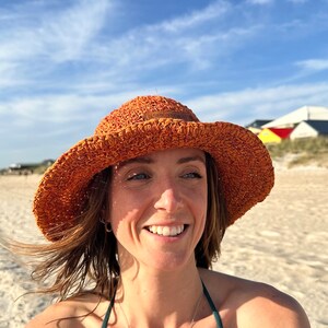 May include: A woman wearing a woven orange sun hat with a wide brim, a dark green bikini top, and a silver necklace. The background shows a beach with white sand and a blue sky.