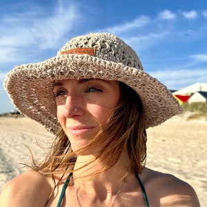 Può includere: Una donna indossa un cappello da sole in crochet beige con un'etichetta in pelle marrone che dice "Spicy Sols" e una collana d'argento con un ciondolo rettangolare. Indossa un top di bikini verde e ha i capelli al vento.