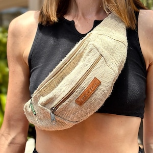 May include: A woman wearing a black tank top and a pleated black skirt is wearing a beige hemp fanny pack with the text "Spicy Sols" on it.