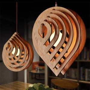 Può includere: Due lampade a sospensione in legno con un design a strati a forma di goccia. Le luci sono realizzate in legno marrone chiaro e hanno un bagliore caldo e invitante. Il design a strati crea un look unico e moderno, perfetto per ogni arredamento.