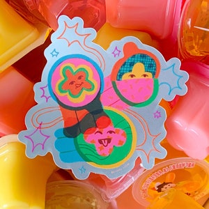 Könnte beinhalten: Ein bunter Aufkleber mit drei Comicfiguren. Die Figuren sind eine rosa Blume mit einem lächelnden Gesicht, eine blaue und rosa Figur mit einem rosa-weiß gestreiften Hut und eine grüne und rosa Figur mit einer rosa Zunge, die herausstreckt. Der Aufkleber befindet sich auf einem Hintergrund aus blauen und weißen Sternen.