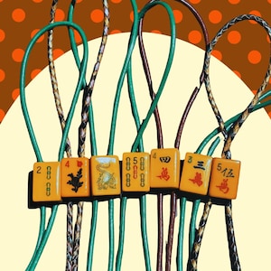 May include: A collection of vintage Mahjong tiles, each with unique symbols and numbers, strung together with green, brown, and braided cords. The tiles are orange with red and black markings. The background is a brown color with orange polka dots.