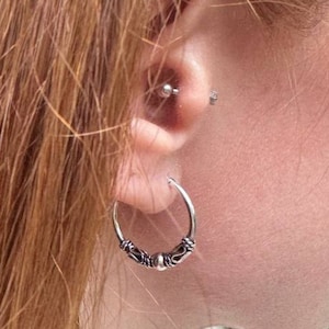 May include: A close-up of a person's ear with a silver hoop earring. The earring has a decorative design with small beads. The person has reddish-brown hair and multiple ear piercings.