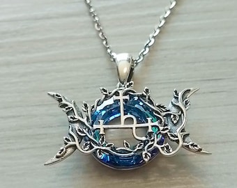 Puede incluir: Collar con colgante plateado con un diseño circular azul y verde, una cruz y otros símbolos. El colgante está enmarcado por una vid frondosa y medias lunas. El collar es de plata.