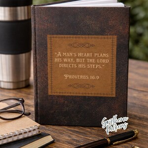 May include: A brown leather-bound journal with the text "A MAN'S HEART PLANS HIS WAY, BUT THE LORD DIRECTS HIS STEPS." PROVERBS 16:9. A black pen, glasses, and a travel mug are also on the wooden surface.