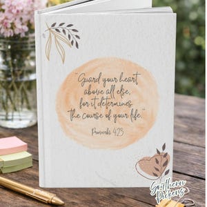 May include: A hardcover journal with the quote "Guard your heart above all else, for it determines the course of your life." in script. The cover features a peach-colored watercolor circle and botanical accents. The journal is on a wooden surface.