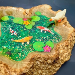 May include: A wooden resin art piece depicting a pond with lily pads, colorful fish, and a white duck. The pond is set in a wooden base with a natural wood grain.